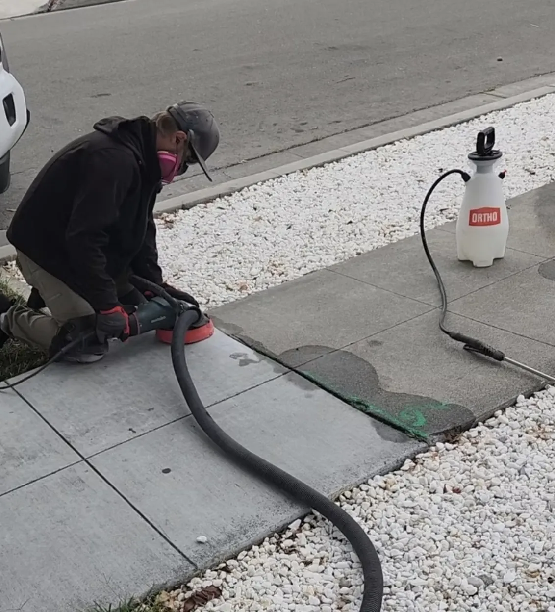 Concrete crew member grinding a new sidewalk edge with a curing sprayer beside white rock